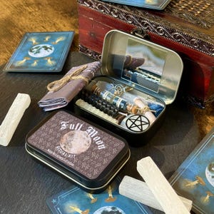 May include: A Full Moon Ritual Kit in a black tin with a full moon graphic. The kit includes a variety of items, such as candles, crystals, and a pentagram. Other items include a small mirror, cards, and a bundle of rolled paper.