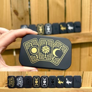 May include: A black metal box featuring a gold design of three tarot cards with celestial symbols. The box is held by a hand, with several other similar boxes visible in the background. The design includes a sun, star, and crescent moon.