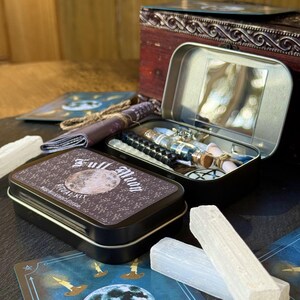 May include: A Full Moon Ritual Kit in a black tin with a full moon design. The kit includes a variety of items, such as candles, crystals, and a small tin with a mirror. Other items include a small scroll, a black beaded bracelet, and tarot cards.