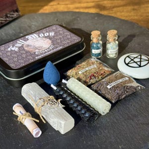 May include: A collection of ritual items including a black tin with "Full Moon Ritual Kit" text, small glass bottles, bags of herbs and rice, candles, a blue incense cone, and a white stone with a pentagram symbol.