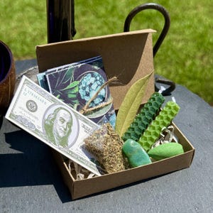 May include: A small, open cardboard box filled with various items. Included are a faux $100 bill, a bundle of dried herbs, green candles, a green stone, a leaf, and a small card with a floral design. The box sits on a dark surface.