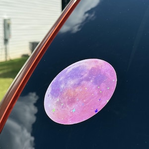 May include: Oval-shaped sticker featuring a pink and purple moon design with star accents. The sticker is affixed to a dark surface, reflecting the sky and clouds. The sticker's design is reminiscent of a celestial theme.