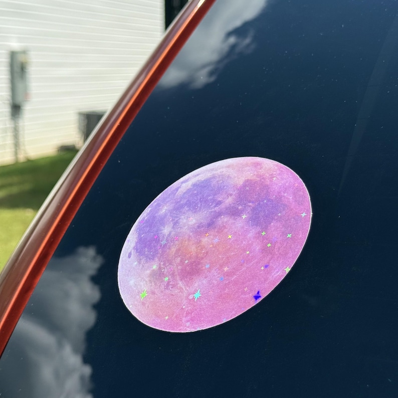 May include: Oval-shaped sticker featuring a pink and purple moon design with star accents. The sticker is affixed to a dark surface, reflecting the sky and clouds. The sticker's design is reminiscent of a celestial theme.