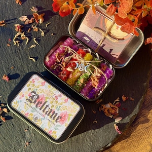 May include: Open metal tin with mini spell candles in various colors, tied with twine and adorned with charms. The tin is labeled "Beltane" with floral accents. Dried flowers and petals surround the tin, creating a mystical aesthetic.