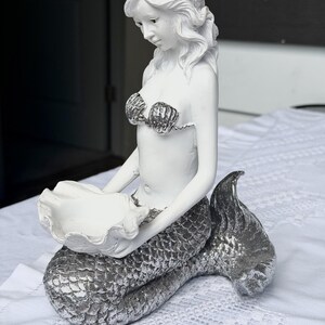 Op de afbeelding: Een wit zeemeerminbeeldje met een zilverkleurige staart en een top in de vorm van schelpen. De zeemeermin zit en houdt met beide handen een grote, open witte schelp vast, die dient als een klein schaaltje voor het bewaren van voorwerpen.