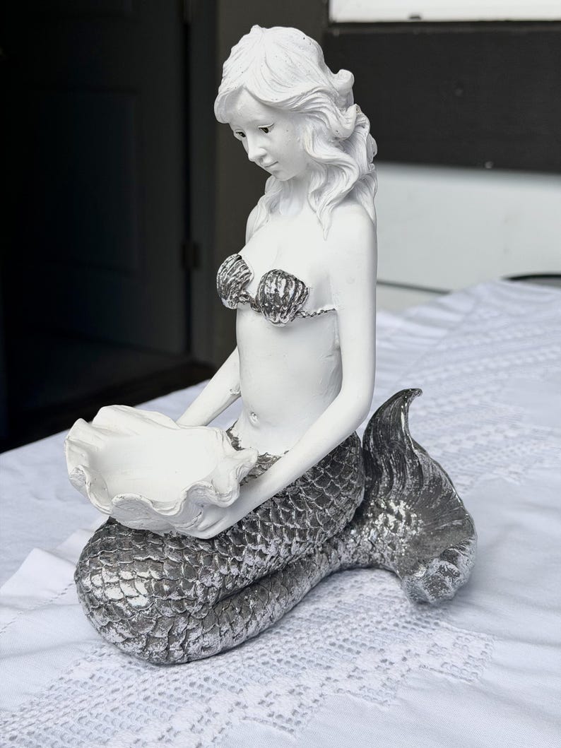 Op de afbeelding: Een wit zeemeerminbeeldje met een zilverkleurige staart en een top in de vorm van schelpen. De zeemeermin zit en houdt met beide handen een grote, open witte schelp vast, die dient als een klein schaaltje voor het bewaren van voorwerpen.