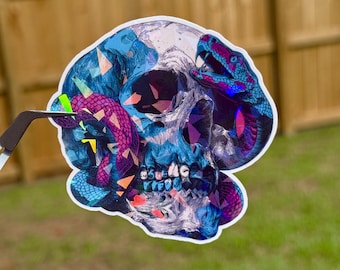 May include: A sticker featuring a skull design with a snake wrapped around it. The skull is primarily blue with purple and white accents. The snake is purple and pink with blue highlights. The sticker is on a green grassy background.