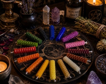 May include: A collection of twelve small, cylindrical beeswax candles with a textured honeycomb pattern, arranged in a circular formation on a black circular board featuring gold celestial illustrations and zodiac symbols.