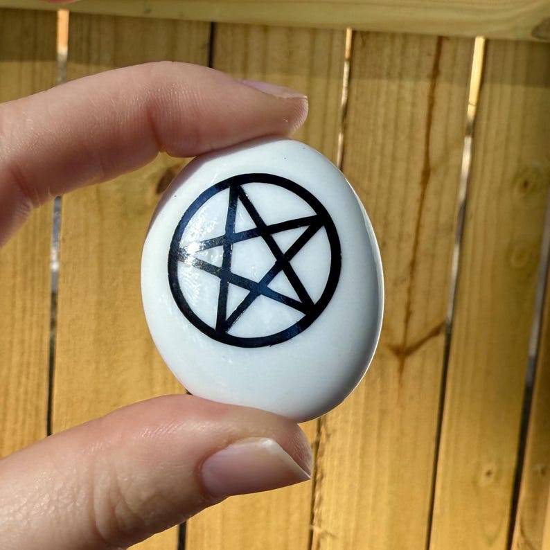 May include: A white, smooth stone with a black pentagram design. The pentagram is enclosed in a black circle. The stone is held up against a wooden fence.
