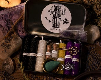 Puede incluir: Una caja metálica rectangular que contiene varias velas de cera de abeja en negro, blanco, amarillo y morado, cada una con una etiqueta. Incluye dos pequeños frascos de vidrio, un incienso en cono verde y dijes en tono dorado y plateado.