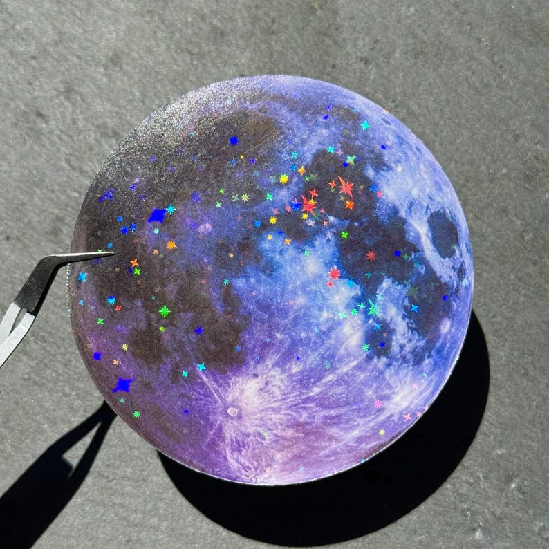 May include: A round sticker featuring a detailed illustration of the moon in shades of purple and blue, with sparkling star accents in various colors. The sticker is being held by tweezers, casting a shadow on a gray surface.