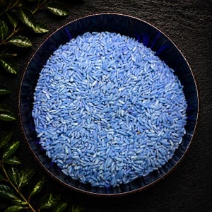 May include: A round, dark blue ceramic bowl filled with a large portion of light blue, long-grain rice. The bowl has a smooth, reflective interior surface and is positioned against a dark, textured background with green leaves visible on the side.