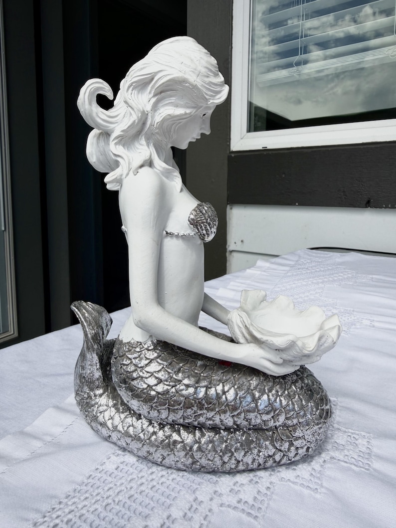 Op de afbeelding: Een wit zeemeerminbeeldje met een metallic zilveren staart en haar. De zeemeermin draagt een schelpvormige top en houdt een grote, open witte schelp in beide handen, met gedetailleerde schubben op de staart.