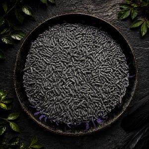 May include: A round, dark-colored pan filled with a dense layer of small, elongated, charcoal-gray grains. The grains are uniform in shape and texture, covering the entire surface of the pan, which features a weathered, dark metallic rim.