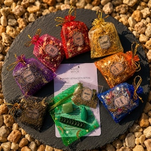 May include: A collection of seven small organza drawstring bags filled with dried herbs and flowers, each with a label. A central white card titled Money Attraction Ritual includes a green bag with a green stone and a small green candle.