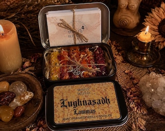 May include: A black metal tin containing mini dressed spell candles in yellow, orange, and red, tied with twine and a silver charm. The lid features a label reading White Witch Lughnasadh Lammas Mini Dressed Spell Candles, with a matching card inside.