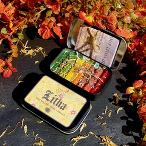 May include: Two open metal tins with mini spell candles. One tin contains green, yellow, orange, and red candles. The other tin has a label that reads "OUT LITHA (SU". A third tin with the text "WHITE WITCH Litha MINI DRESSED SPELL CANDLES" is also visible.
