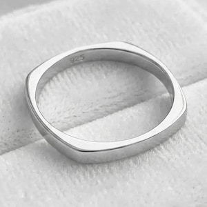 May include: A polished silver ring with a square shape and rounded corners. The ring is marked with "925" on the inside. The ring is displayed on a white, textured surface.