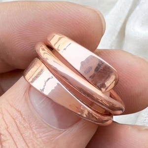 May include: A set of three rose gold rings. Each ring has a flat, rectangular design. The rings are stacked together, creating a layered effect. The metal has a polished, reflective surface.