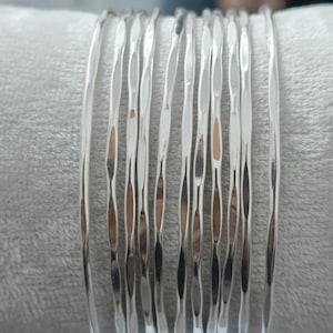 Op de afbeelding: Een stapel zilverkleurige armbanden met een getextureerd, gehamerd ontwerp. De armbanden zijn op elkaar gestapeld, waardoor ze licht reflecteren en een glinsterend effect creëren. De armbanden liggen op een zachte, grijze ondergrond.