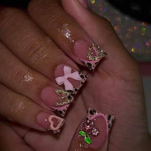 May include: A close-up of a hand with long, decorated acrylic nails. The nails are primarily pink with leopard print accents. Some nails feature gold embellishments, bows, and heart designs. The overall style is glamorous and feminine.