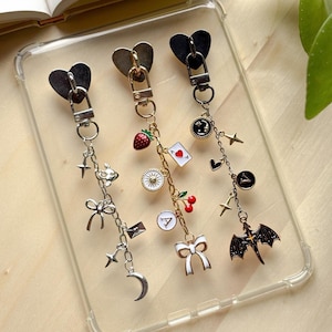 May include: Three decorative keychains with heart-shaped clasps. One keychain is silver with charms including a crescent moon, stars, and a bow. Another is gold with a strawberry, cherries, and a bow. The third is black with a dragon charm.
