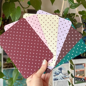 Könnte beinhalten: Eine Hand hält einen Fächer aus bunten Karten. Jede Karte hat eine abgerundete Oberseite und ist in einer anderen Farbe gehalten, darunter Burgunderrot, Rosa, Gelb und Grün. Jede Karte ist mit kleinen weißen Punkten bedeckt. Die Karten werden vor einem Hintergrund aus grünen Pflanzen präsentiert.