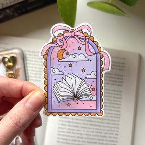 Könnte beinhalten: Ein Aufkleber mit einem fantasievollen Design eines offenen Buches in einem Fensterrahmen. Der Rahmen ist mit einer rosa Schleife, Wolken, Sternen und einem Mond in Pastelltönen verziert. Der Aufkleber hat einen dekorativen, wellenförmigen Rand.