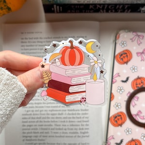 Pode incluir: Um adesivo de acrílico transparente com uma pilha de quatro livros em tons de rosa e laranja, encimada por uma abóbora laranja. O design inclui uma vela rosa claro, uma pequena margarida branca, dois cogumelos, uma lua crescente amarela e pequenas estrelas.