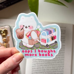 Könnte beinhalten: Ein Aufkleber mit einem weißen Katzencharakter, der einen braunen Eiskaffee und einen Spiegel hält, mit einem Bücherwagen. Der rosa Text lautet "oops I bought more books". Der Aufkleber hat einen hellblauen Hintergrund.