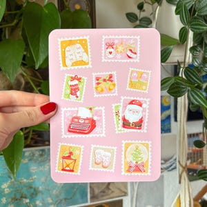 Peut inclure: Une feuille d'autocollants rectangulaire rose avec des illustrations sur le thème de Noël dans des bordures de timbres-poste. Les motifs comprennent un pull, des ornements, une chaussette, des cloches, le Père Noël, une machine à écrire et un sapin de Noël.