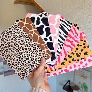 Könnte beinhalten: Eine Hand hält einen Fächer aus gemusterten Karten. Die Karten zeigen verschiedene Tiermuster, darunter Leoparden-, Giraffen-, Kuh- und Zebrastreifen sowie abstrakte Muster in Braun-, Schwarz-, Rosa- und Orangetönen. Die Karten sind rechteckig mit abgerundeten Ecken.