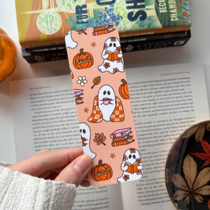 May include: A rectangular bookmark with a light orange background featuring a repeating pattern of white ghosts, orange pumpkins with the word read, stacks of books, and small brown leaves. The ghosts are depicted reading books or wearing blankets.