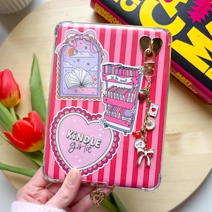 A hand holding a decorated Kindle with a striped pink Kindle insert, bookish stickers, a Kindle chain, and a dust plug charm.