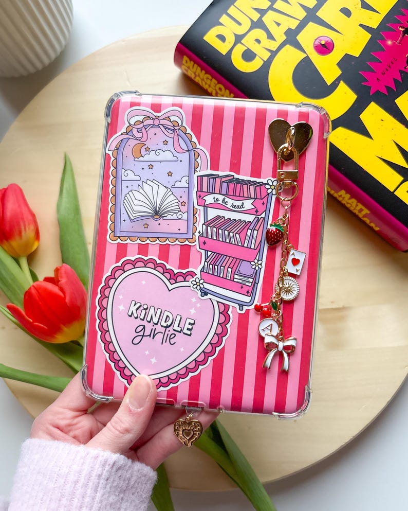 A hand holding a decorated Kindle with a striped pink Kindle insert, bookish stickers, a Kindle chain, and a dust plug charm.