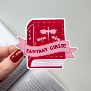 Könnte beinhalten: Ein roter Aufkleber in Form eines Buches mit den Worten "FANTASY GIRLIE" auf einem rosa Banner. Das Buch hat eine Drachenillustration und drei Herzen. Der Aufkleber wird von einer Hand mit rotem Nagellack gehalten.