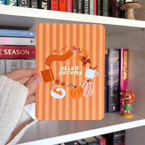 May include: A rectangular print with a vertical orange and light orange striped background featuring the text HELLO AUTUMN in white. Illustrations include a scarf, acorns, a mug, a slice of pie, and two pumpkins in orange and brown tones.