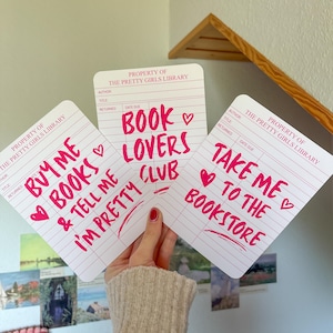 Könnte beinhalten: Drei weiße Karten im Bibliotheksstil mit rotem Text und Herzen. Die Karten lauten "Buy Me Books & Tell Me I'm Pretty", "Book Lovers Club" und "Take Me to the Bookstore". Die Karten werden von einer Person gehalten.