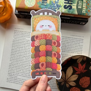 Op de afbeelding: Een gestanste boekenlegger met een wit, rond figuurtje met roze wangen in een bed met een bruin hoofd- en voeteneinde. Het bed is bedekt met een dikke, textuurrijke quilt die bestaat uit vele kleine, kleurrijke, bedrukte cirkelvormige lappen.