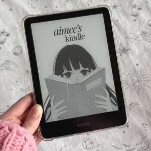 May include: A black Kindle e-reader with a clear protective case displays an illustration of a person reading a book. The screen reads "aimee's kindle" in cursive. The word "kindle" is also printed at the bottom of the screen.