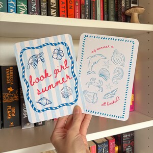 Può includere: Due cartoline rettangolari a tema nautico. La carta a sinistra presenta strisce azzurre, un bordo a corda blu, una tartaruga marina, un ibisco e conchiglie con il testo rosso "book girl summer". La carta a destra mostra illustrazioni marine blu e il testo rosso "my summer is all booked!".