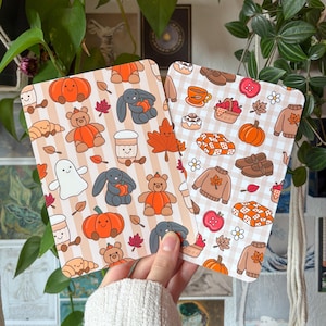 Puede incluir: Dos tarjetas rectangulares con motivos otoñales. La tarjeta izquierda tiene un fondo de rayas beige con ilustraciones de calabazas, ositos, conejos, fantasmas y hojas. La tarjeta derecha tiene un patrón de cuadros vichy blancos con jerséis, cestas de manzanas, mantas y zapatos.