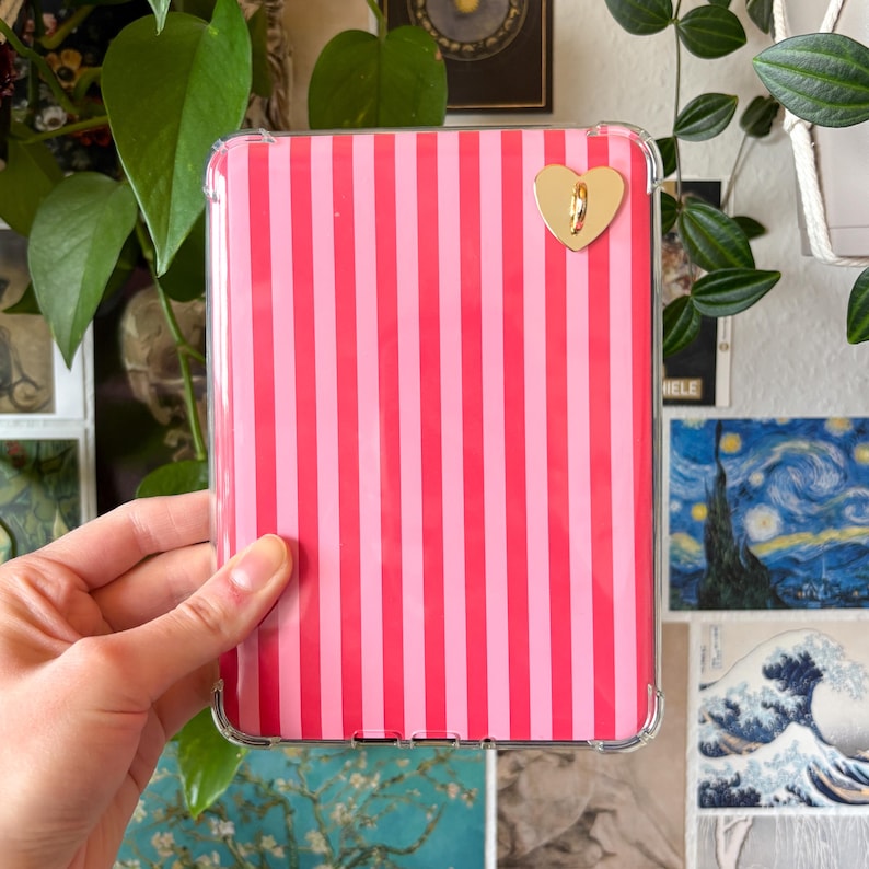 May include: A pink and light pink striped tablet case with a gold heart-shaped clasp. The case is held in a clear protective cover. The background includes green plants and framed artwork.