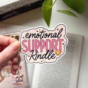 Könnte beinhalten: Ein gestanzter Aufkleber mit den Worten "emotional SUPPORT Kindle" in einer verspielten Schriftart. Der Aufkleber hat einen hellpfirsichfarbenen Hintergrund mit rosa und schwarzem Text und einem kleinen gelben Smiley. Der Aufkleber wird von einer Hand gehalten.