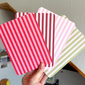 Könnte beinhalten: Eine Hand hält eine Sammlung farbenfroher Karten mit Streifen. Die Karten weisen vertikale Streifen in Rot-, Rosa-, Weiß- und Grüntönen auf. Die Karten haben abgerundete Ecken.