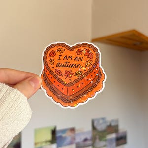 Könnte beinhalten: Ein herzförmiger Aufkleber mit einem orangefarbenen Kuchen, braunen Frosting-Rändern und verstreuten braunen Herbstblättern. Der Text "I AM AN autumn" steht in schwarzer Schreibschrift in der Mitte. Der Aufkleber hat einen holografischen Sterneffekt.