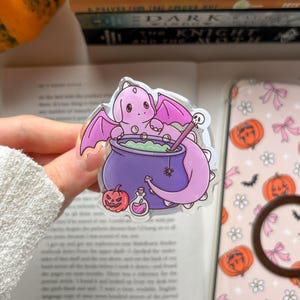 Könnte beinhalten: Eine durchsichtige Acryl-Anstecknadel mit einem hellvioletten Drachen mit Flügeln hinter einem violetten Kessel mit grüner Flüssigkeit. Das Design zeigt einen kleinen Rührstab mit Totenkopf, eine winzige Spinne, einen Kürbis und eine kleine Flasche.