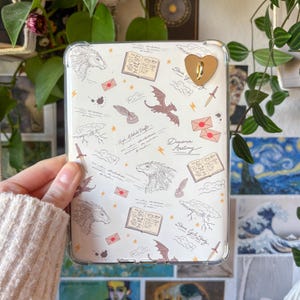 Op de afbeelding: Een doorzichtige tablethoes met een witte achtergrond met een fantasiethema. Het ontwerp bevat illustraties van draken, boeken, enveloppen en tekst. Een goudkleurig hartvormig accessoire is aan de hoes bevestigd.