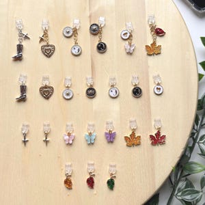 May include: A selection of clip-on earrings featuring charms like a cowboy boot, heart, sun, moon, butterfly, and maple leaf. The earrings are silver and gold, with colourful enamel details. They are presented on a wooden surface.