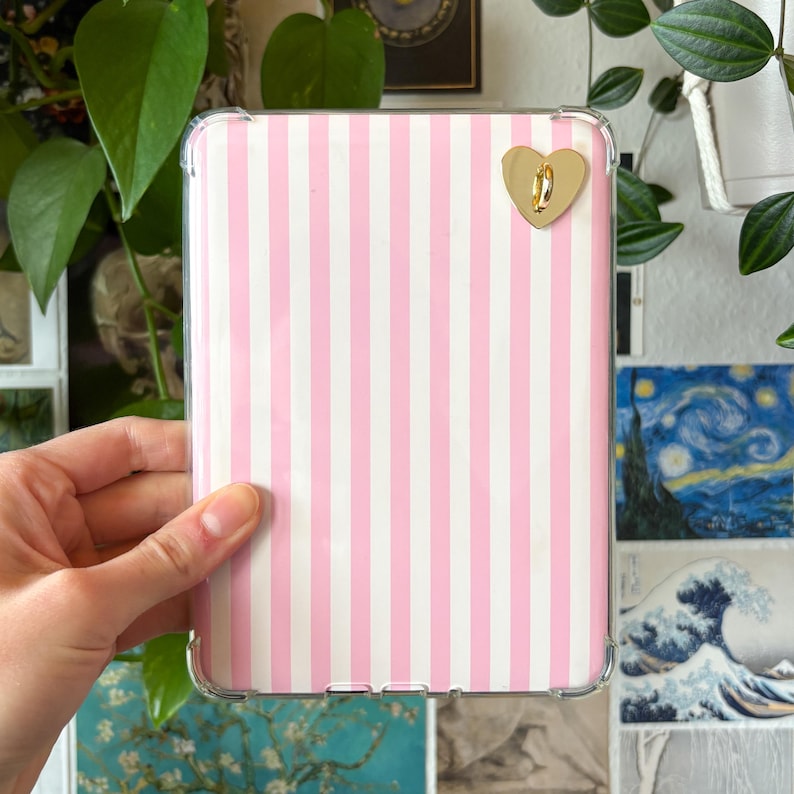 May include: A clear tablet case with a pink and white striped pattern. A gold heart-shaped charm is attached to the case. The case is held in a person's hand.